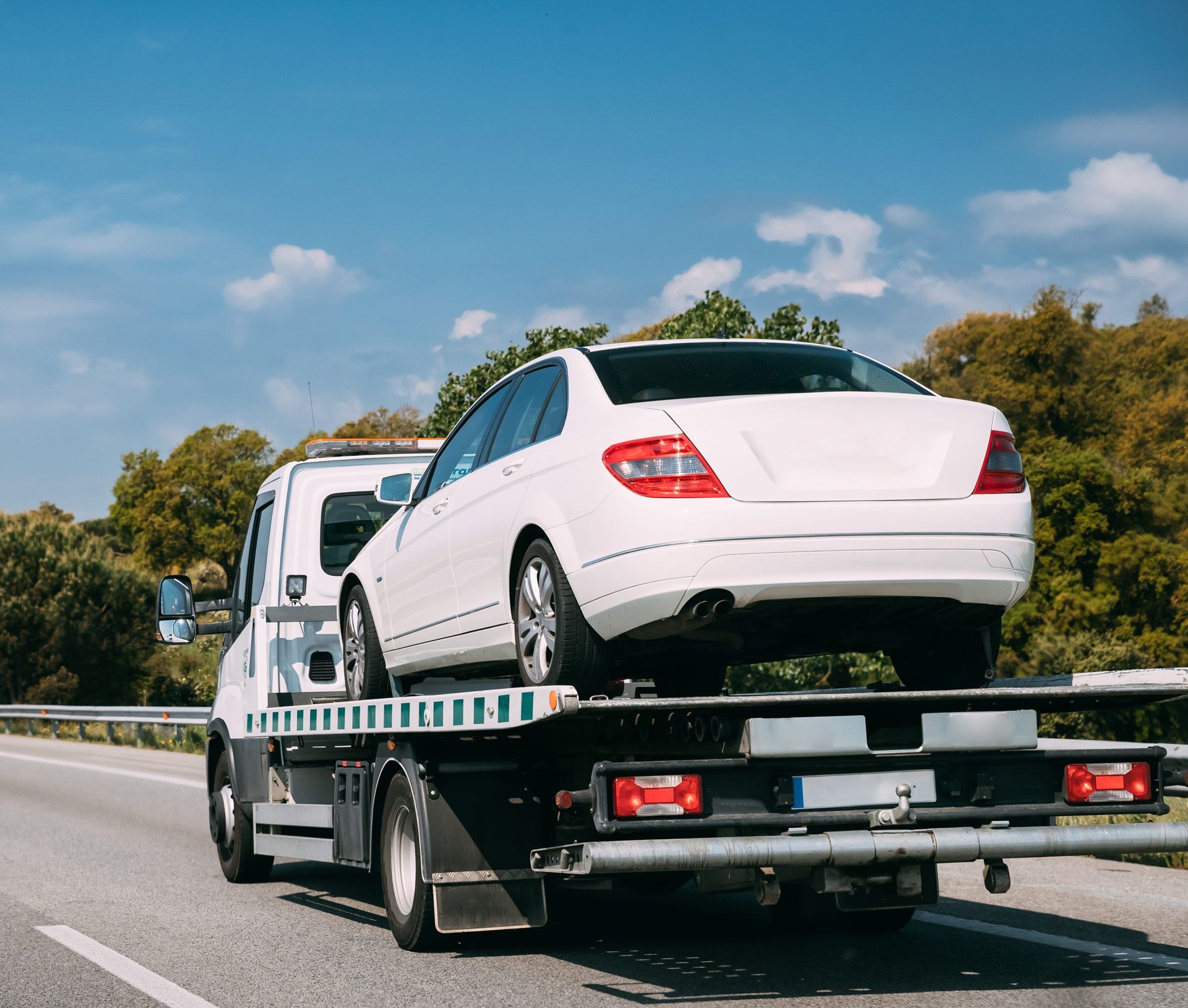 White sedan being towed on a flatbed tow truck on a highway on a sunny day.