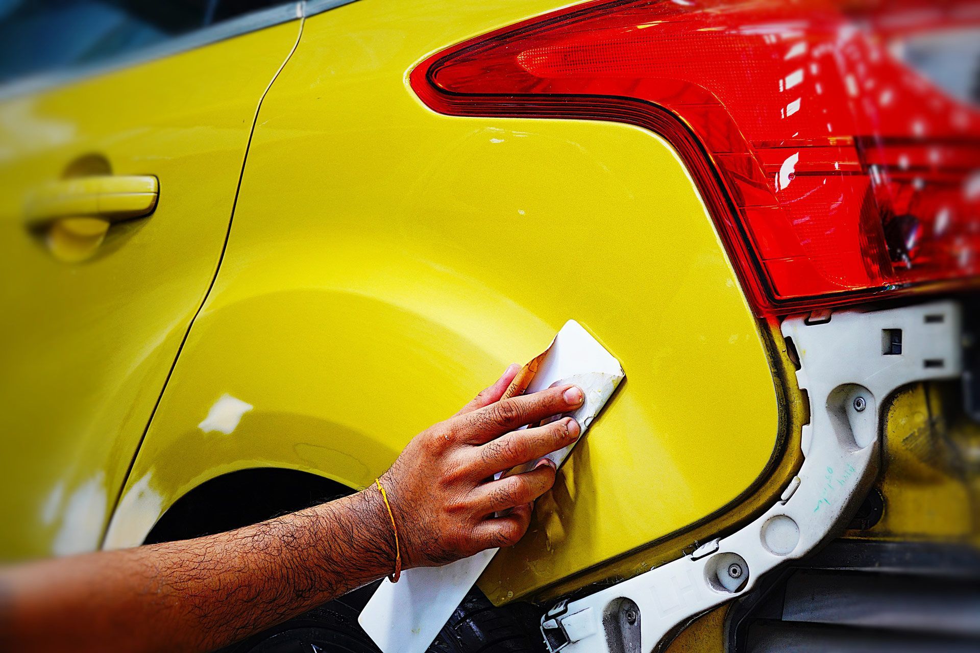 Hand wiping a yellow car's rear quarter panel, near a red taillight and exposed bumper.