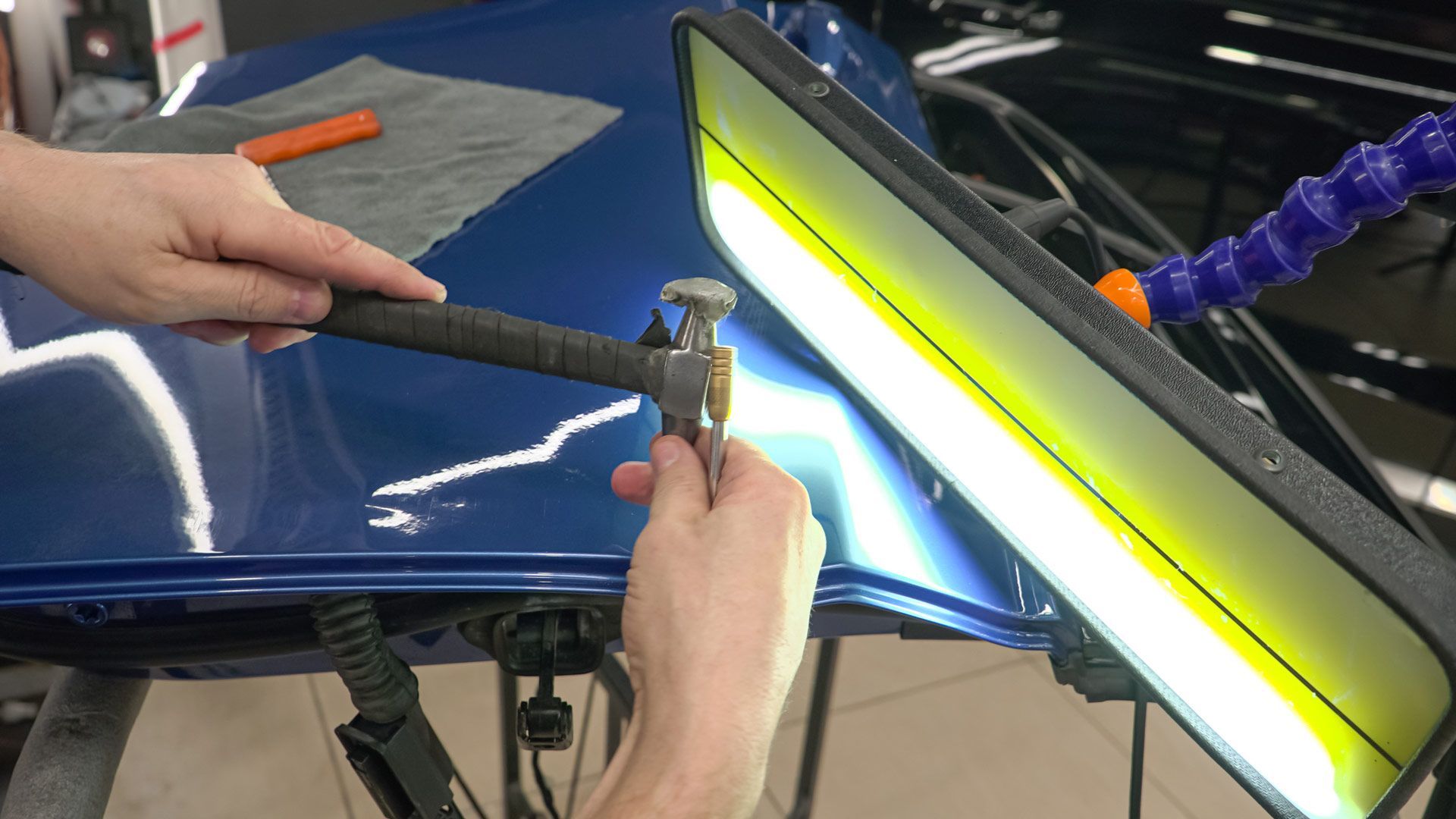 Hands using a hammer to repair a dent in a blue car panel, illuminated by a reflective light.