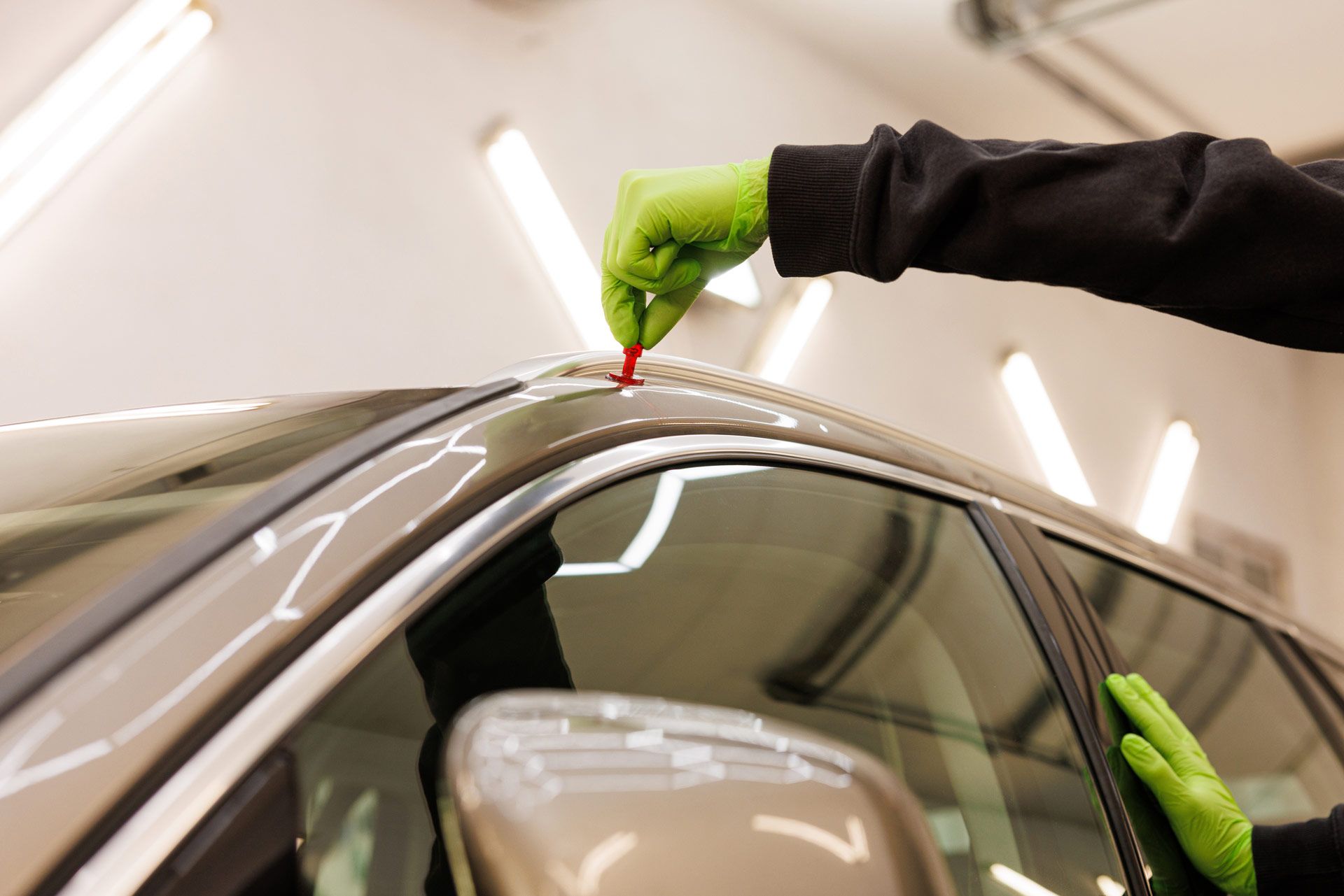 Person in green gloves uses a tool on a car's roof, likely for detailing in a brightly lit garage.