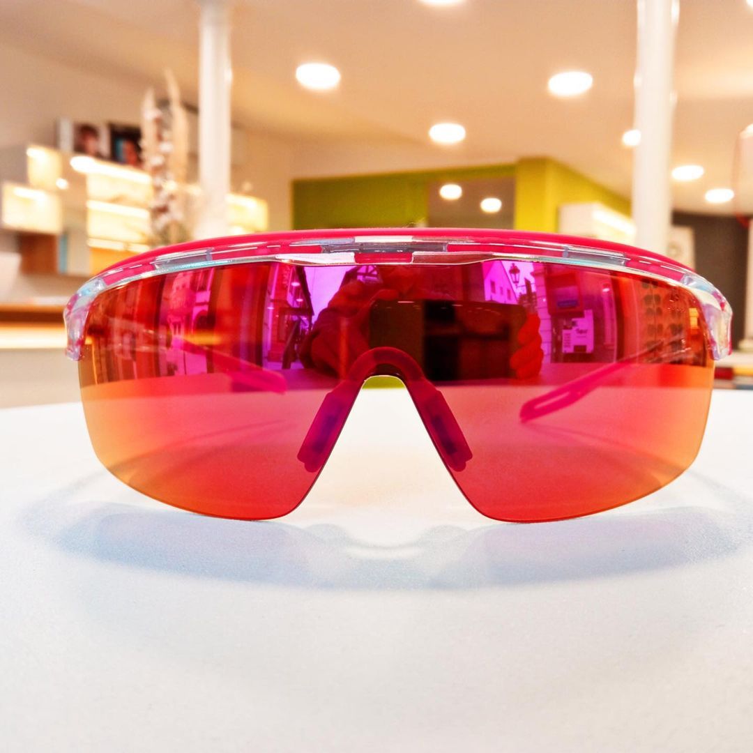 Sonnenbrille mit rosa Rand und reflektierenden roten Gläsern auf einer weißen Oberfläche.