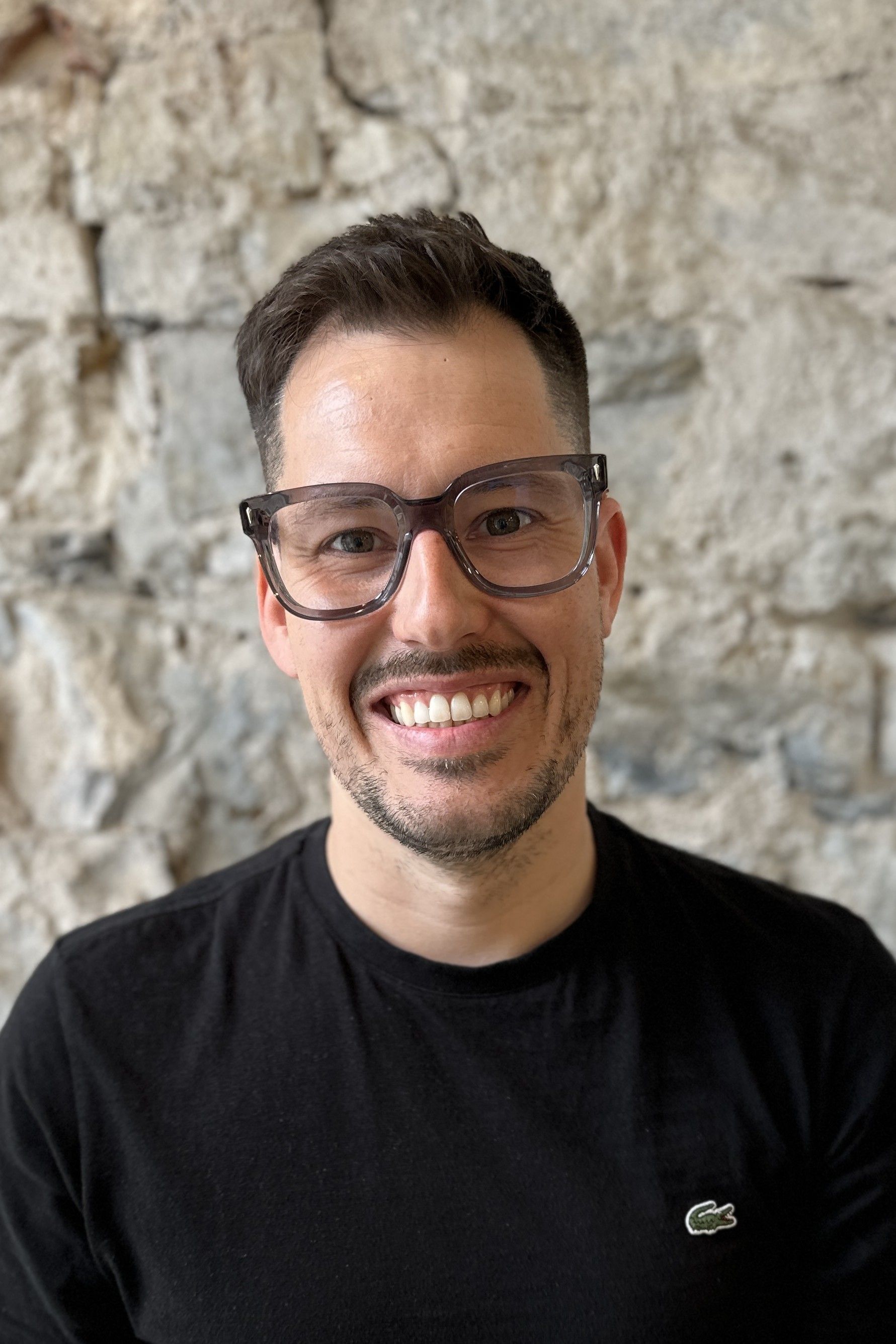 Mann mit Brille lächelt vor einer Steinmauer in die Kamera.