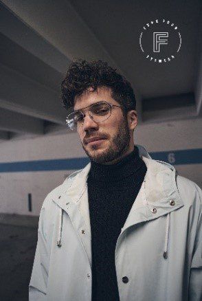 Mann mit lockigem Haar, Brille und Rollkragenpullover, trägt einen hellen Mantel. Steht in einem Parkhaus aus Beton.