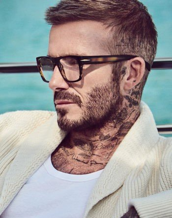 David Beckham mit Brille, Tattoos und weißem Pullover vor türkisfarbenem Hintergrund.