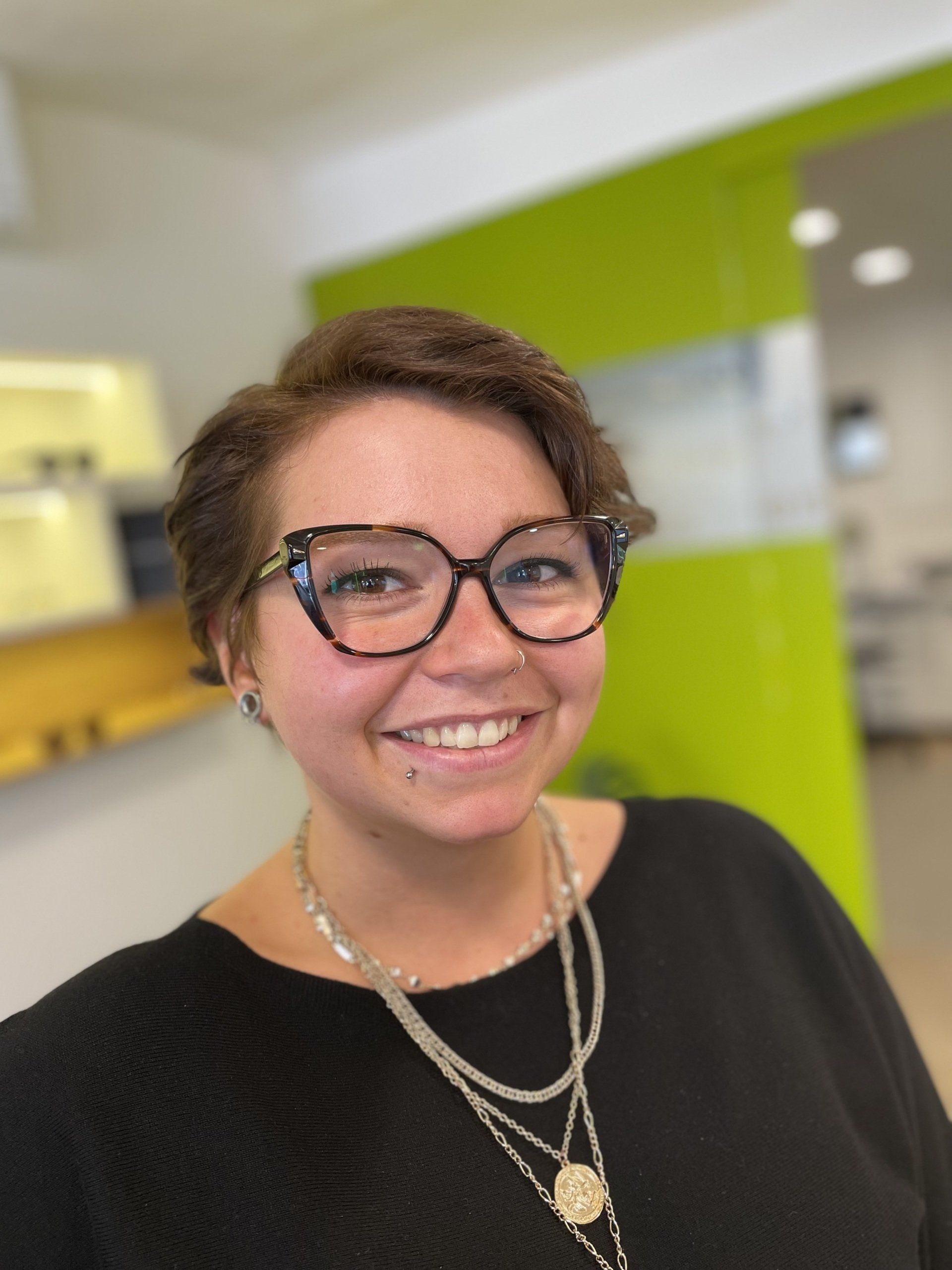 Frau mit kurzen braunen Haaren, Brille, lächelnd. In einem Augenoptikergeschäft.