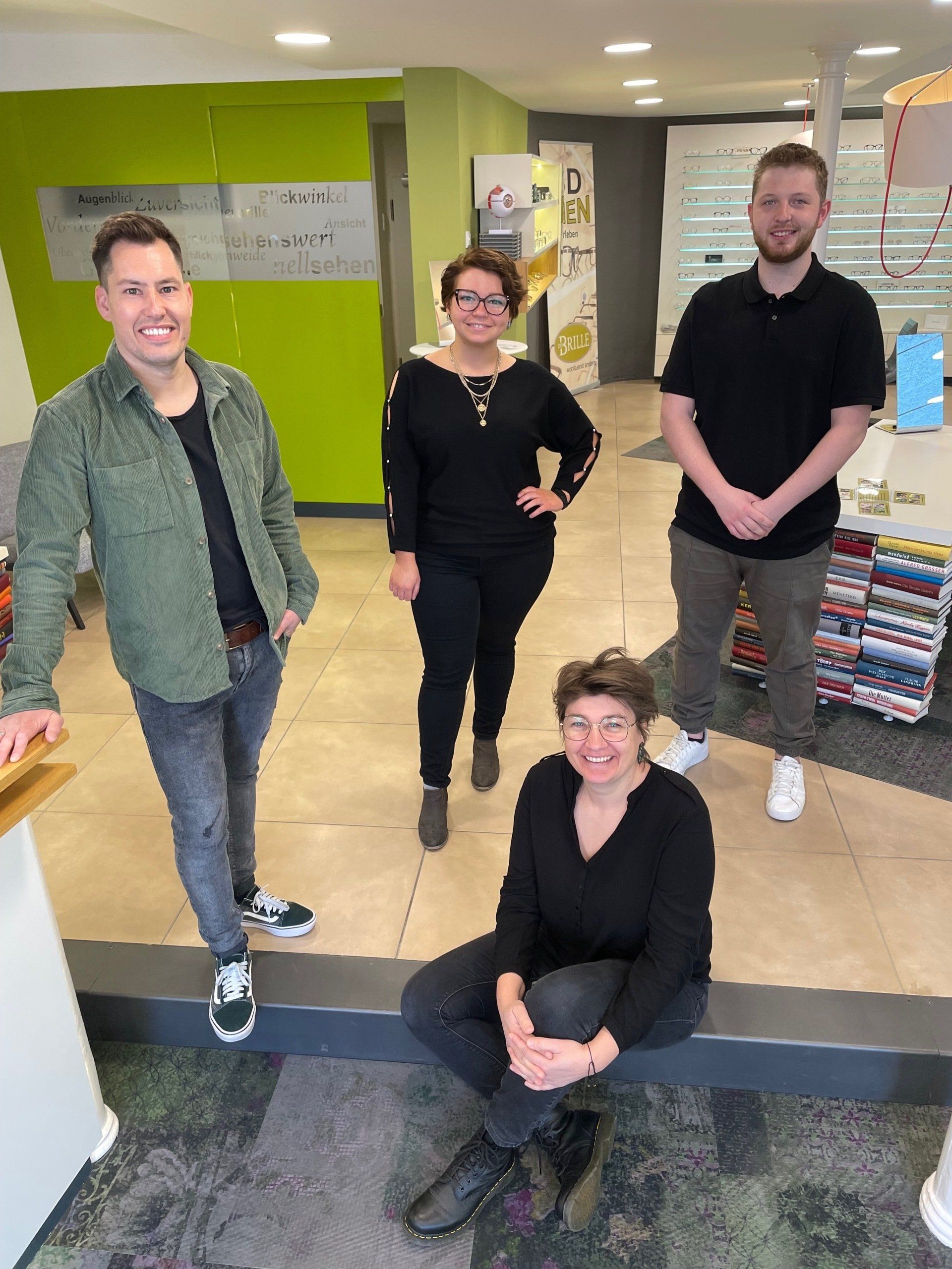 Vier Personen in einem Büro. Zwei Männer und zwei Frauen. Eine Frau sitzt, die anderen stehen.