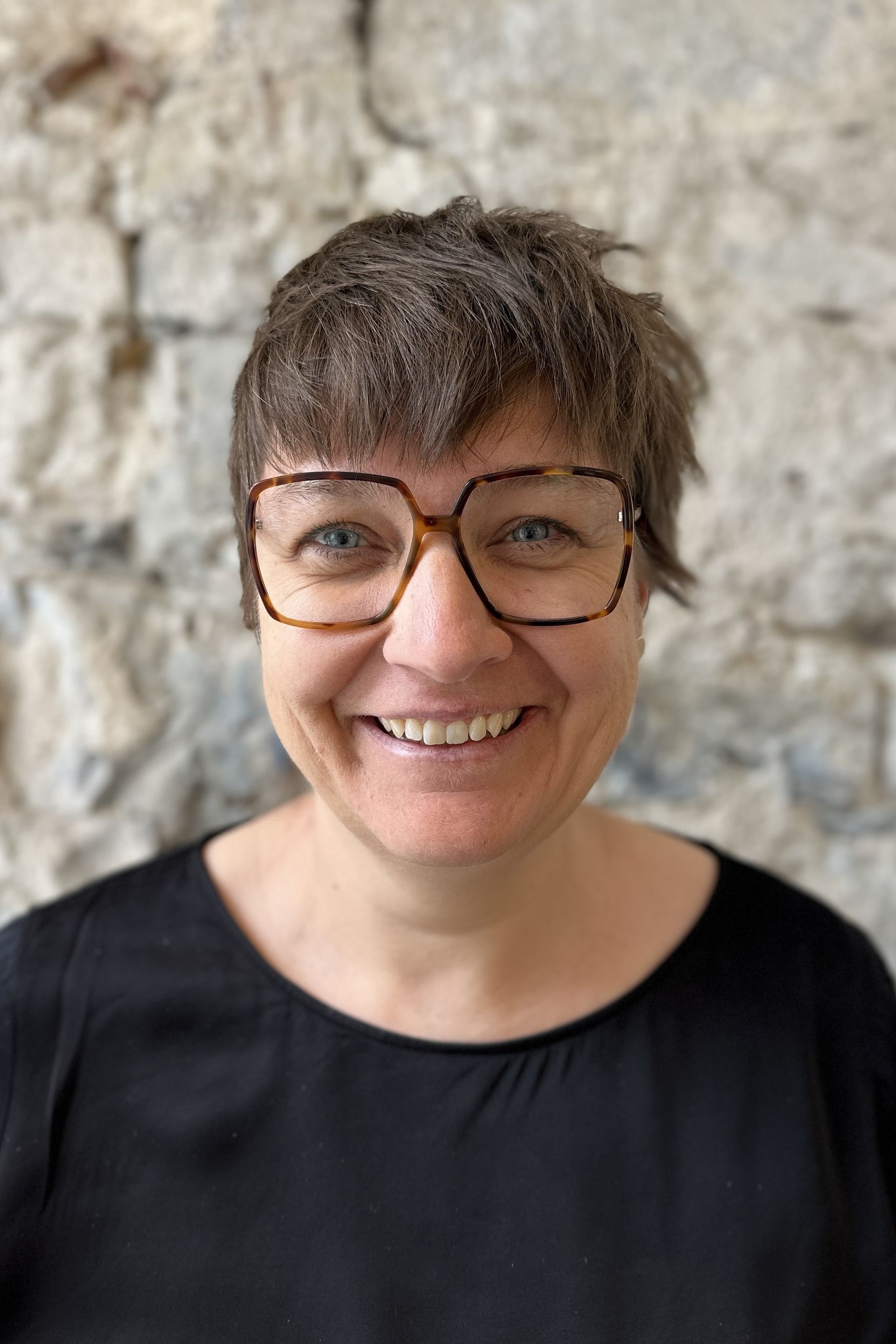 Frau mit kurzen braunen Haaren und Brille lächelt vor einer Steinmauer.