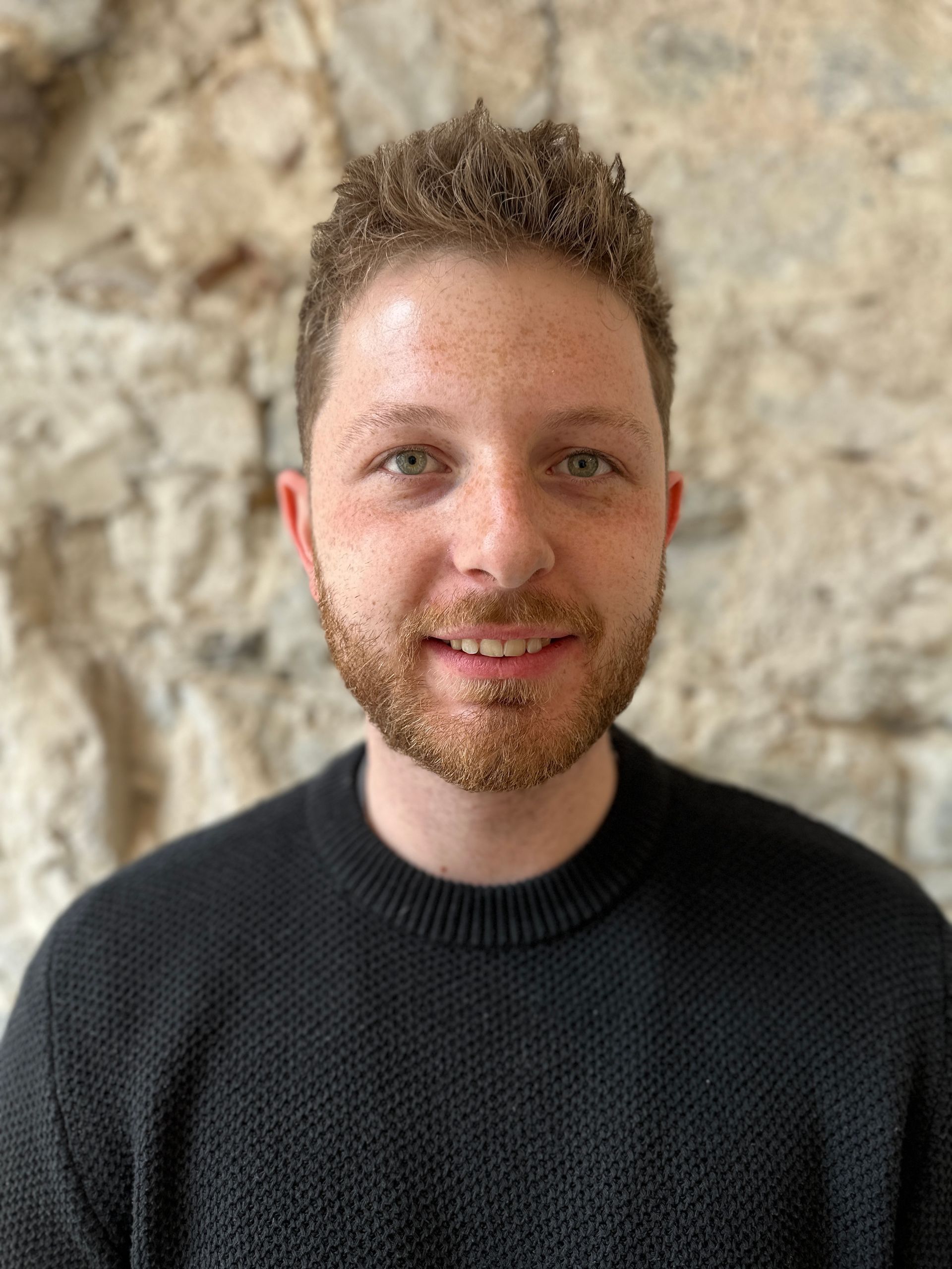 Mann mit lockigem braunem Haar und Bart lächelt vor einer Steinmauer und trägt einen schwarzen Pullover.