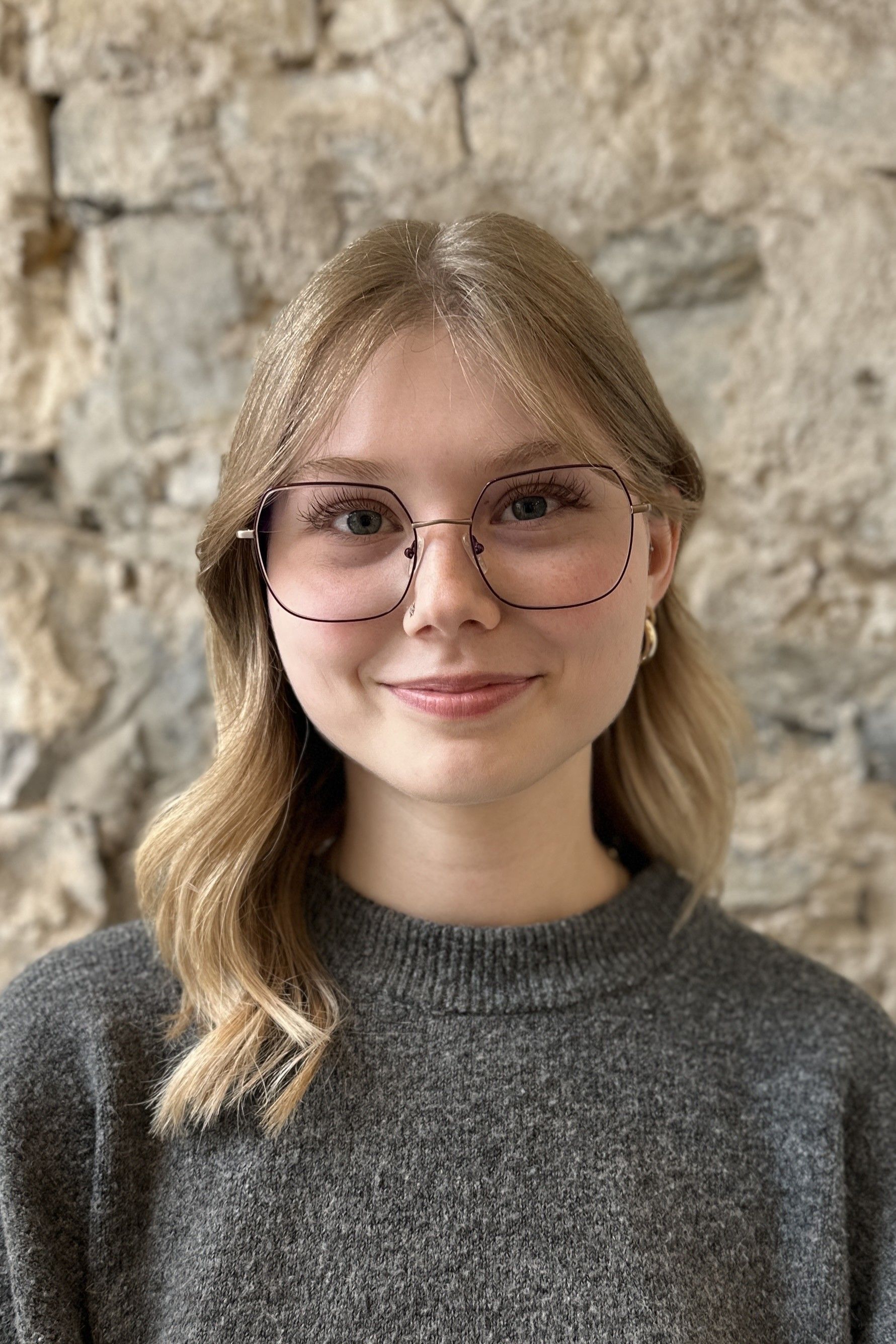 Frau mit blonden Haaren und Brille lächelt vor einer Steinmauer.
