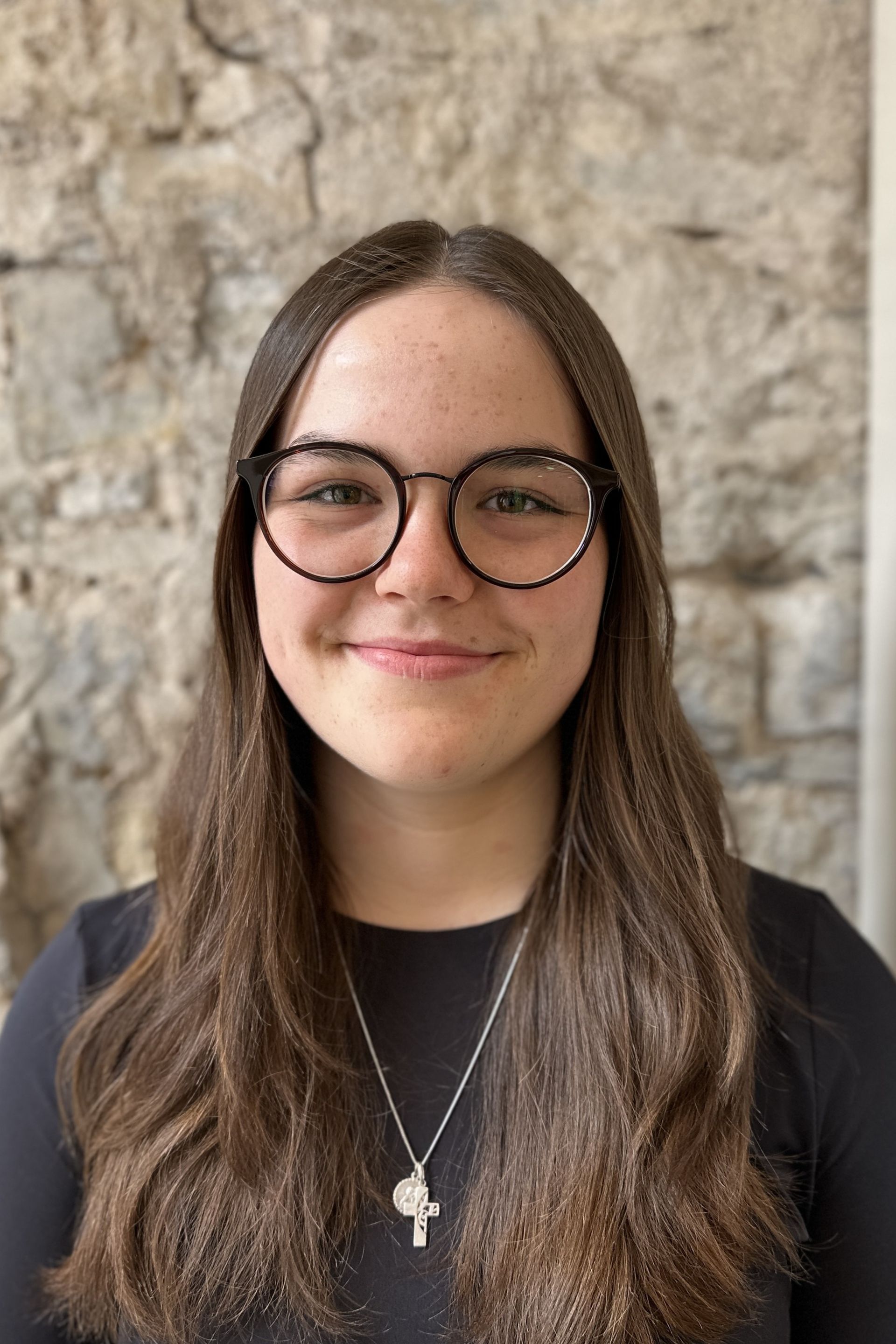 Frau mit langen braunen Haaren und Brille lächelt vor einer Steinmauer.