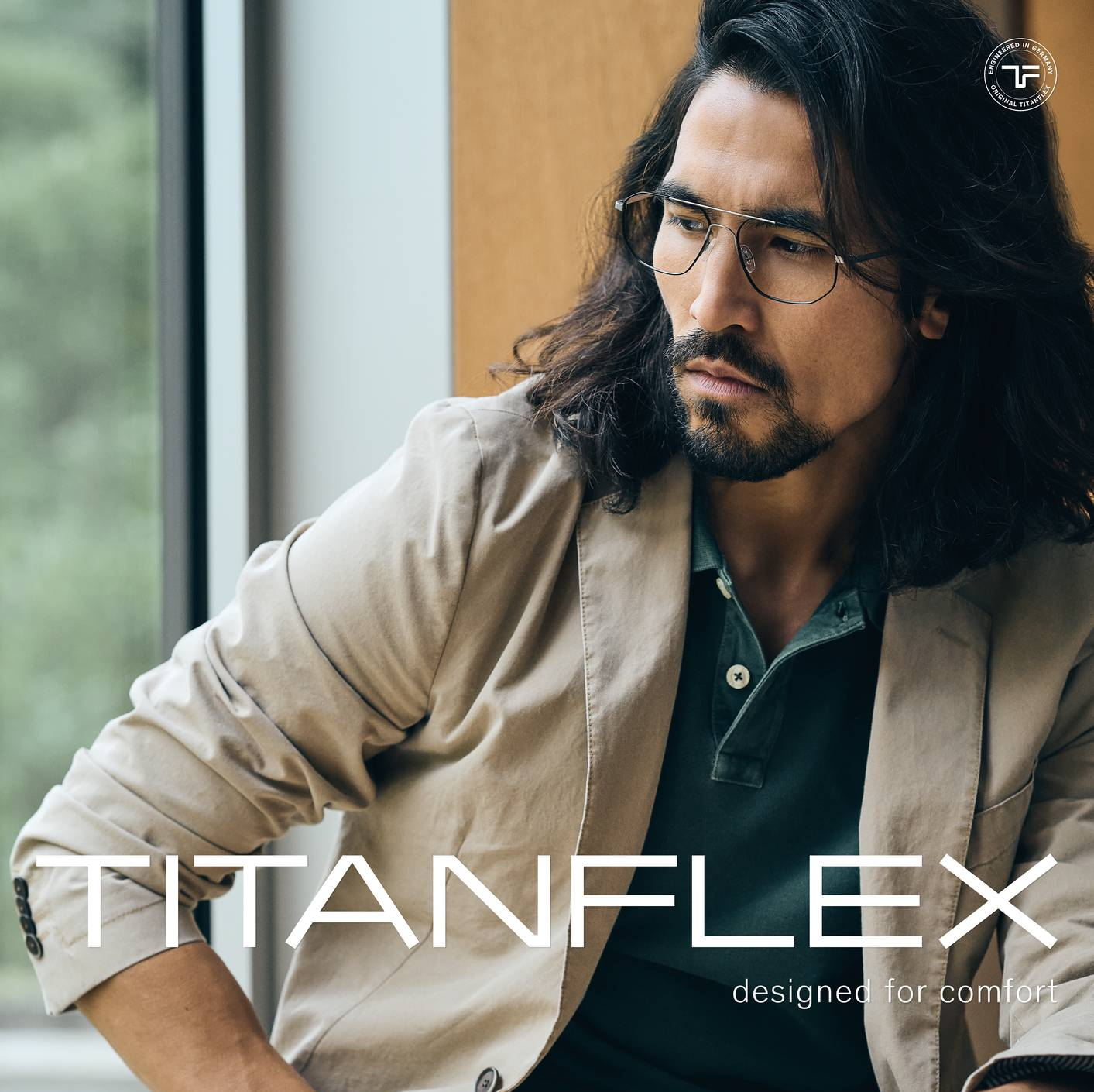 Mann mit Brille sitzt drinnen, trägt beigen Blazer und grünes Poloshirt. „Titanflex“-Logo.