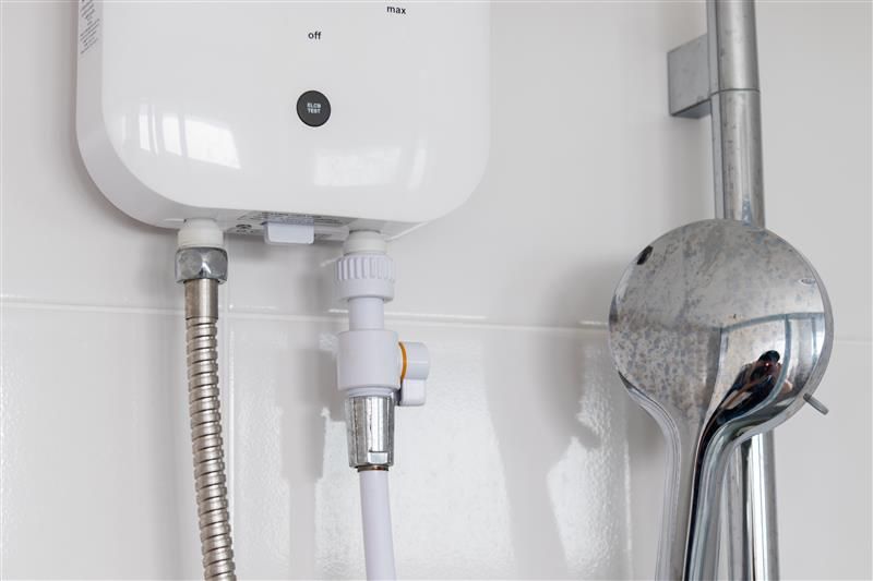 White electric water heater mounted on a white tiled wall next to a shower head.