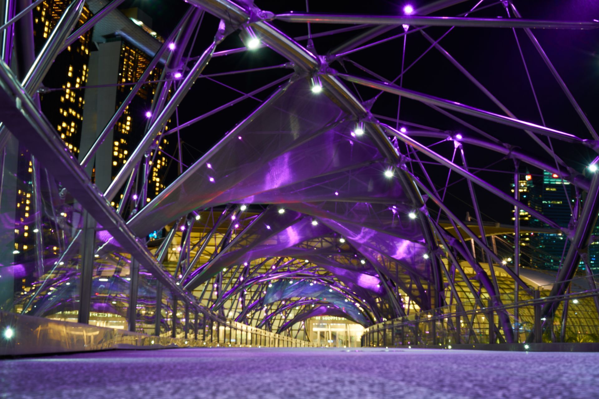 Purple-lit modern pedestrian bridge at night with steel arches and city lights in the background
