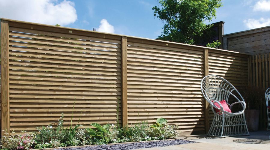 A modern horizontal slat wooden fence creates a privacy screen in a sunny garden, with a white chair on a patio nearby.
