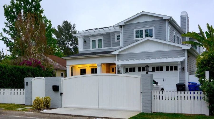 A two-story grey house with white trim and a white picket fence, viewed from the street behind a large white privacy gate.