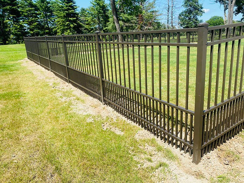 Brown metal fence bordering a grassy area with trees in the background under a blue sky.