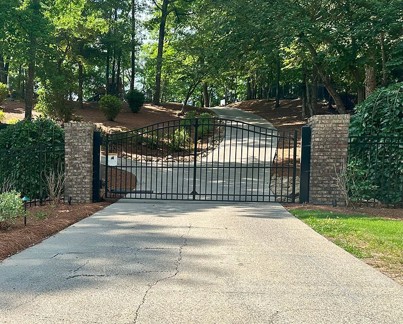 Driveway with closed black metal gate between brick columns, leading to a tree-lined hill.