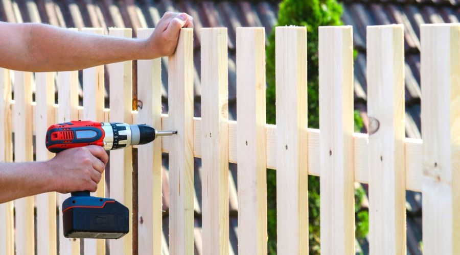 Handyman using a cordless drill to install wooden pickets on a garden fence.