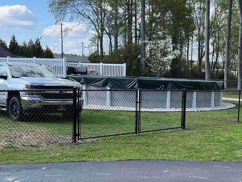 Silver truck beside an above-ground pool with a dark cover, enclosed by chain link and white fences.