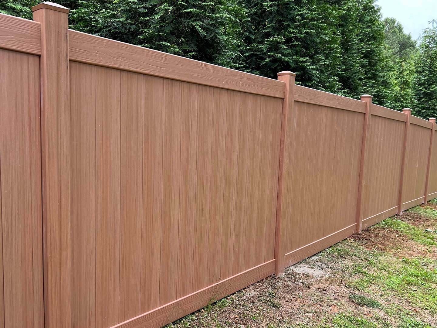 Brown wood-look fence with vertical panels and posts, in front of a grassy area and trees.