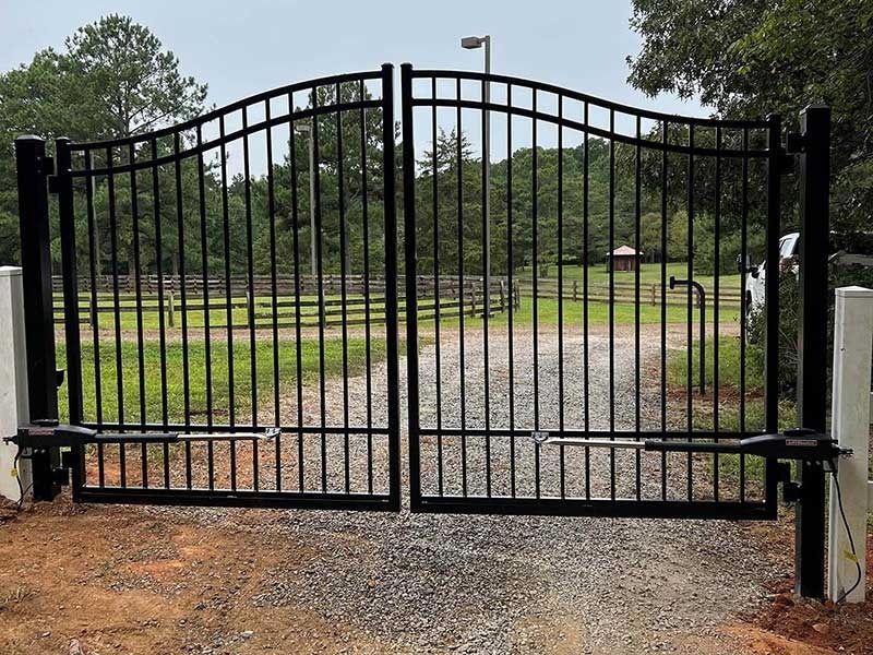 Black metal driveway gate, arched top, opens to gravel road. Green trees in background.