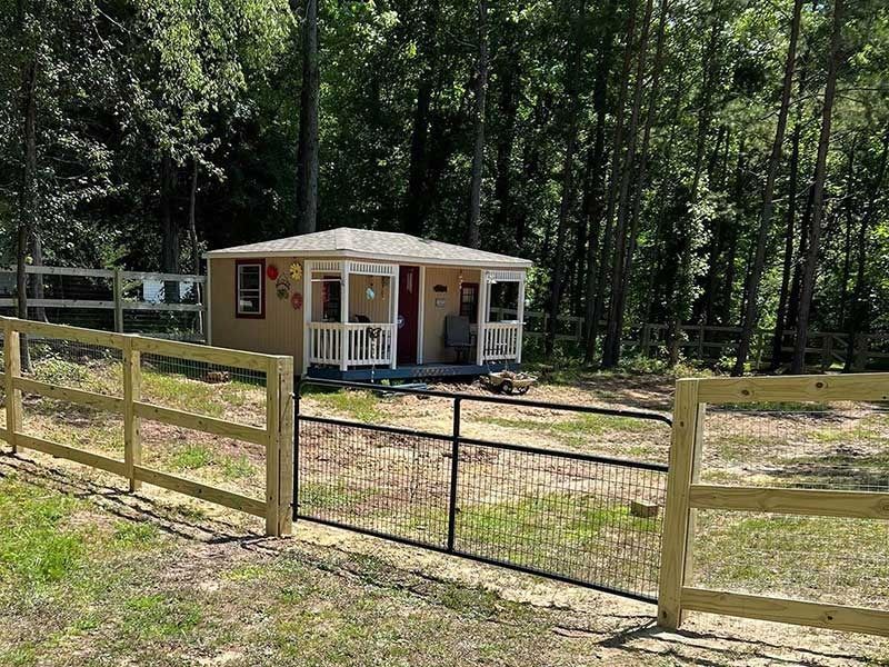 Tan cabin with porch, surrounded by a new wood fence and a black metal gate, set in a wooded area.