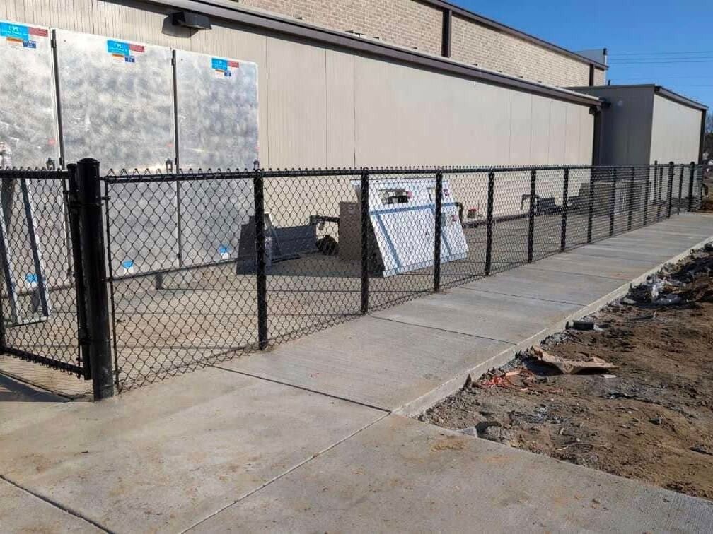 Black chain-link fence along a concrete walkway in front of a tan building.
