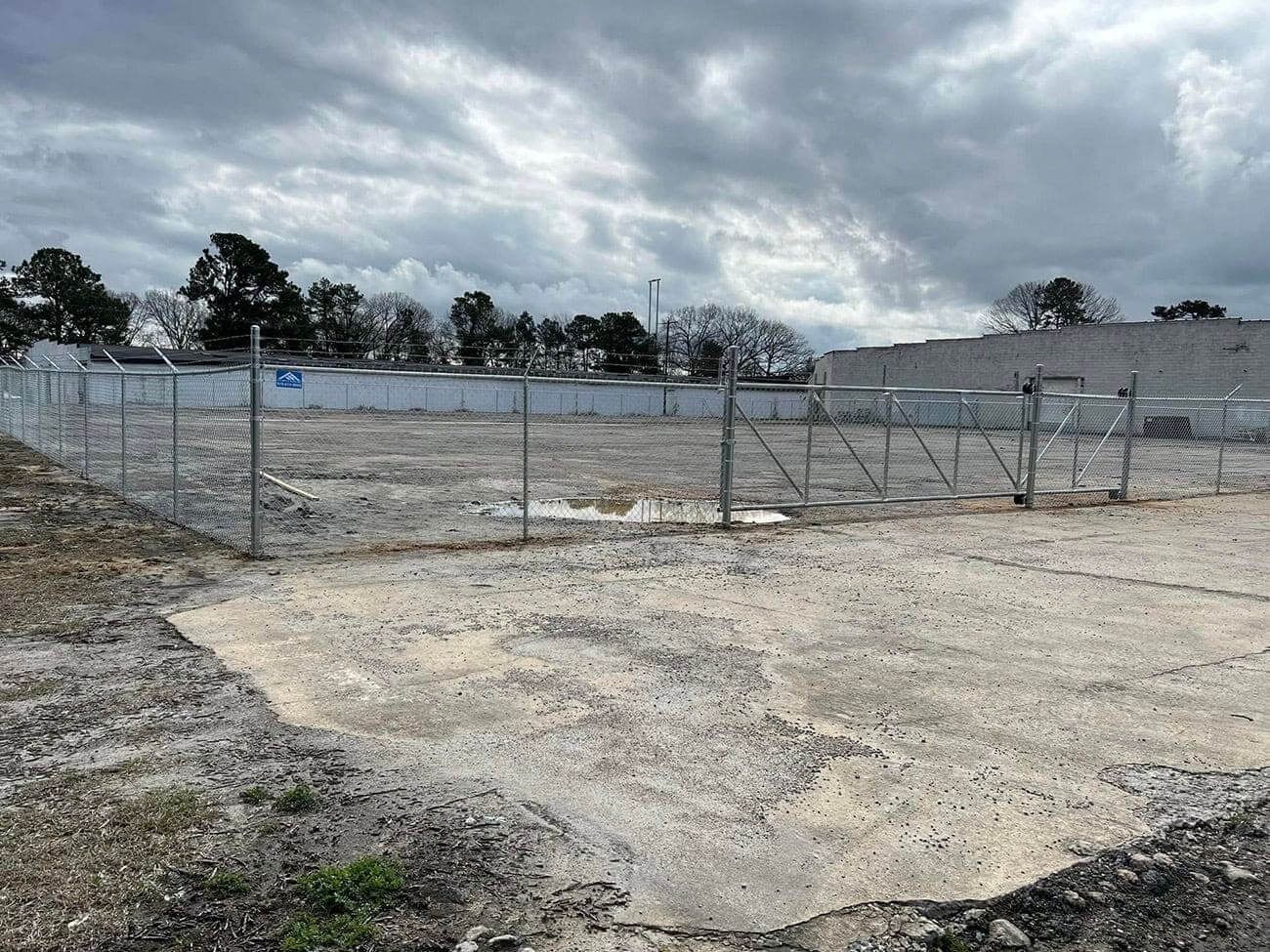 Fenced gravel lot under overcast sky, surrounded by trees.