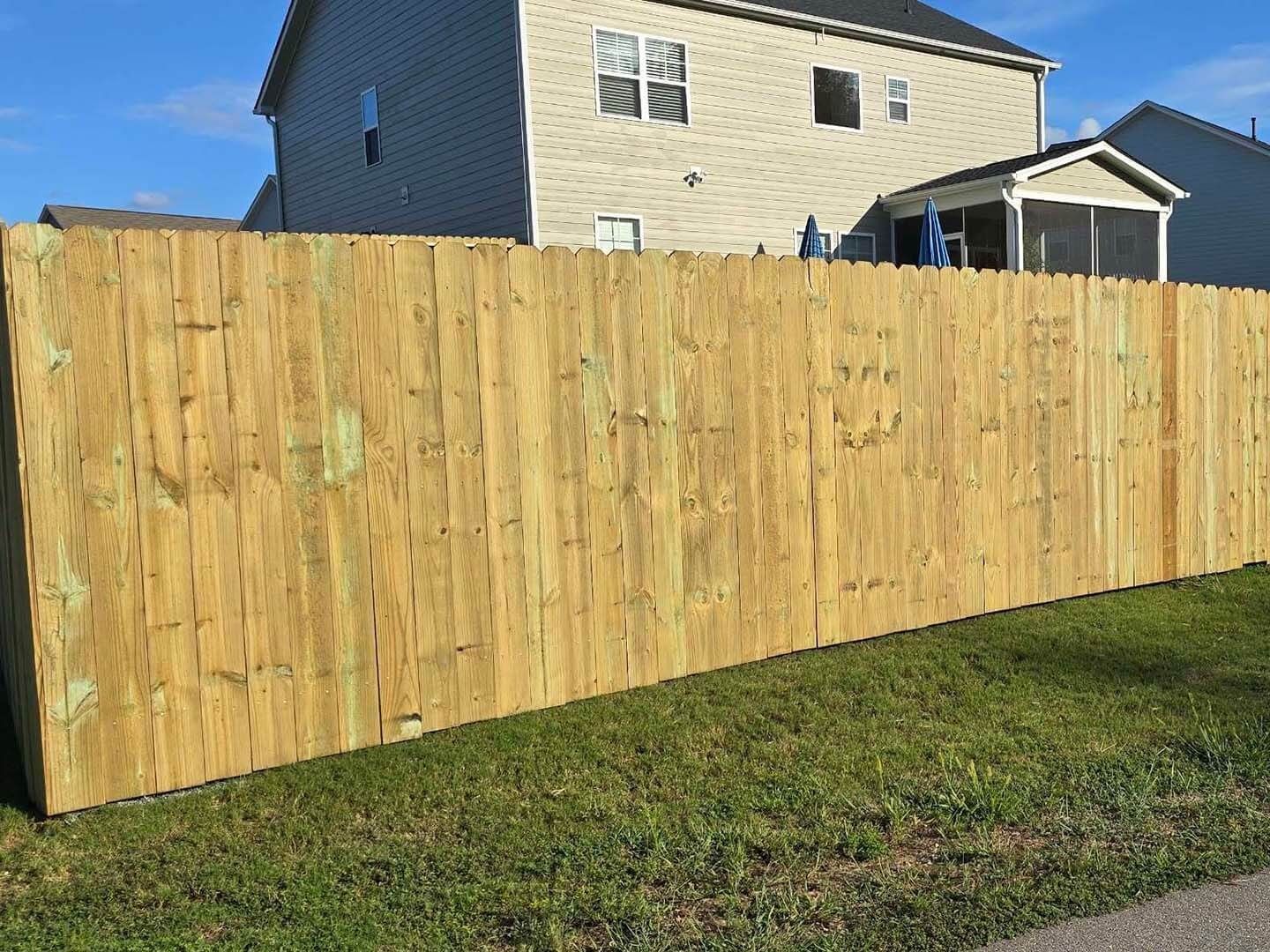 Wooden fence in front of a beige house, on green grass under a blue sky.