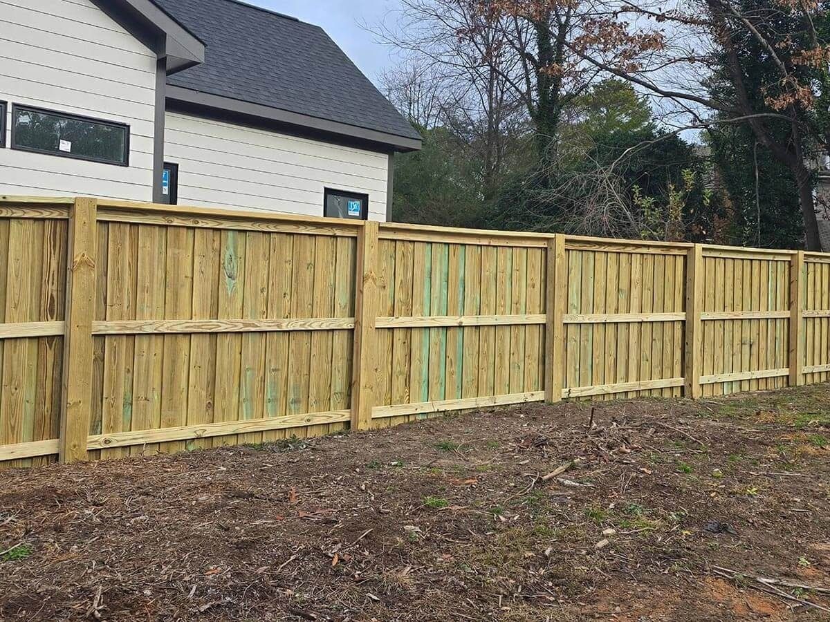 Wooden fence in front of a house with a gray roof. Dry grass and trees in the background.