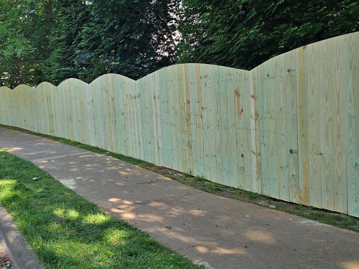 Green wooden fence with wavy top next to a paved walkway and grassy area. Trees in background.