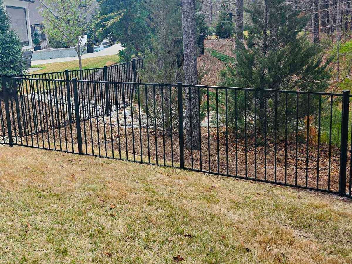 Black metal fence surrounding a grassy backyard with trees and a house in the distance.