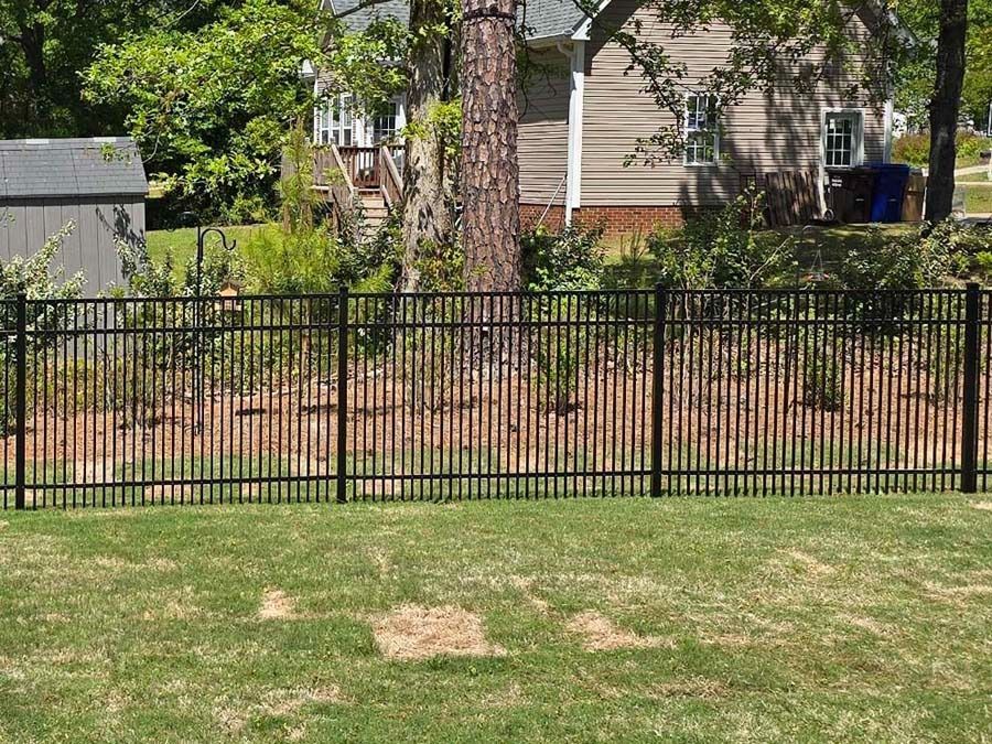 Black metal fence in front of a grassy yard, with a house and trees in the background.