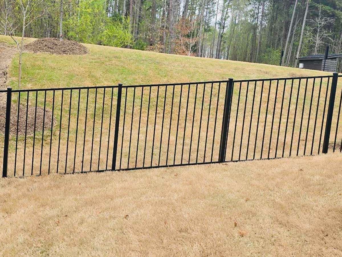 Black metal fence in front of a grassy yard, with a hill and trees in the background.