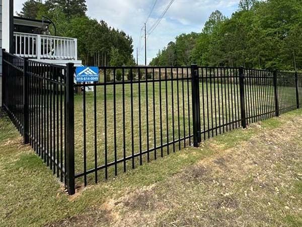 Black metal fence encloses a grassy yard next to a house with trees in the background.