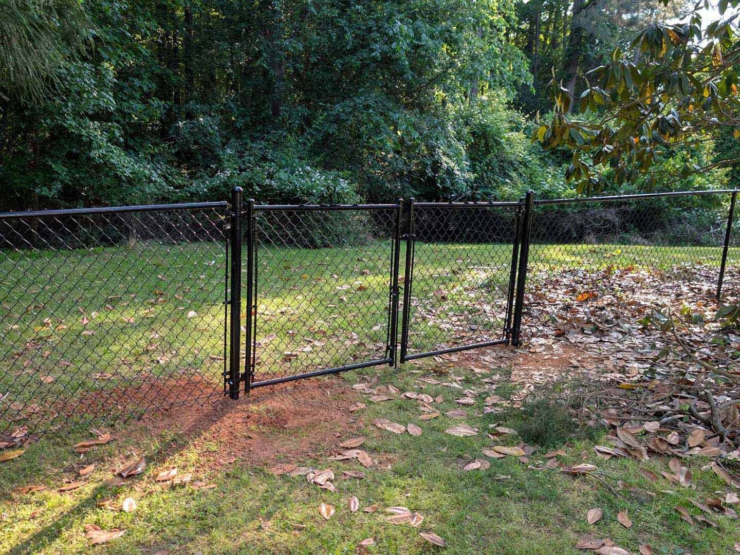 Black chain-link fence with a closed gate in a grassy yard, trees in the background.