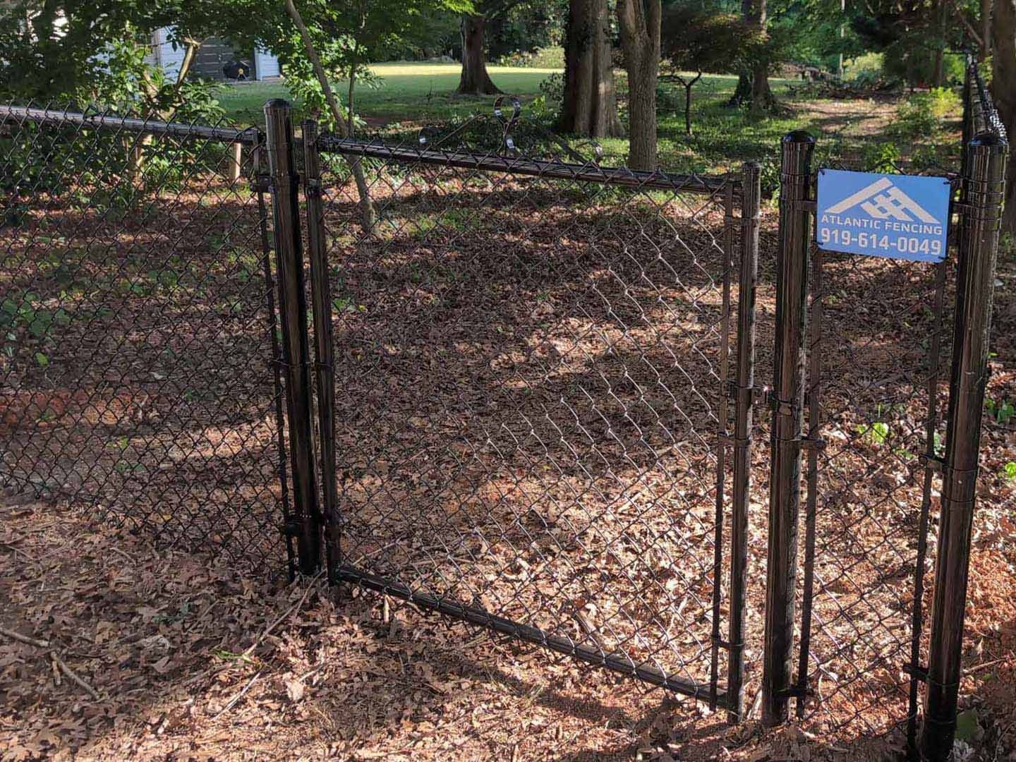 Black chain-link fence gate, open, in a leaf-covered yard with a blue sign on the right post.