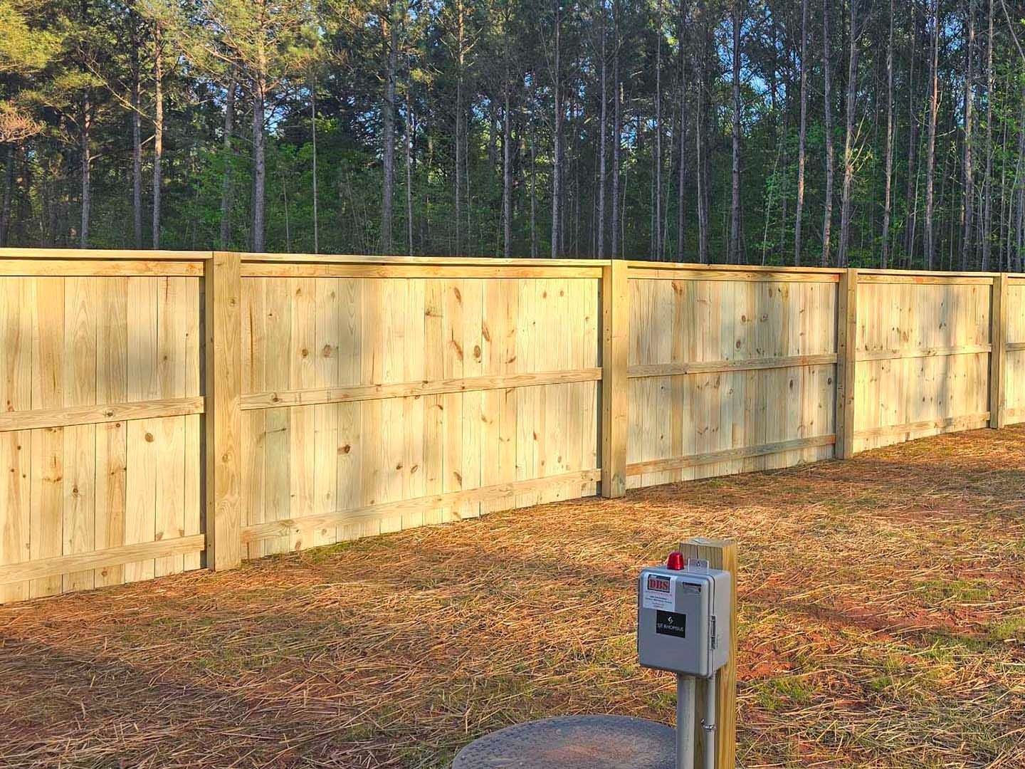 Wooden privacy fence in a yard with a forest background and a utility box.