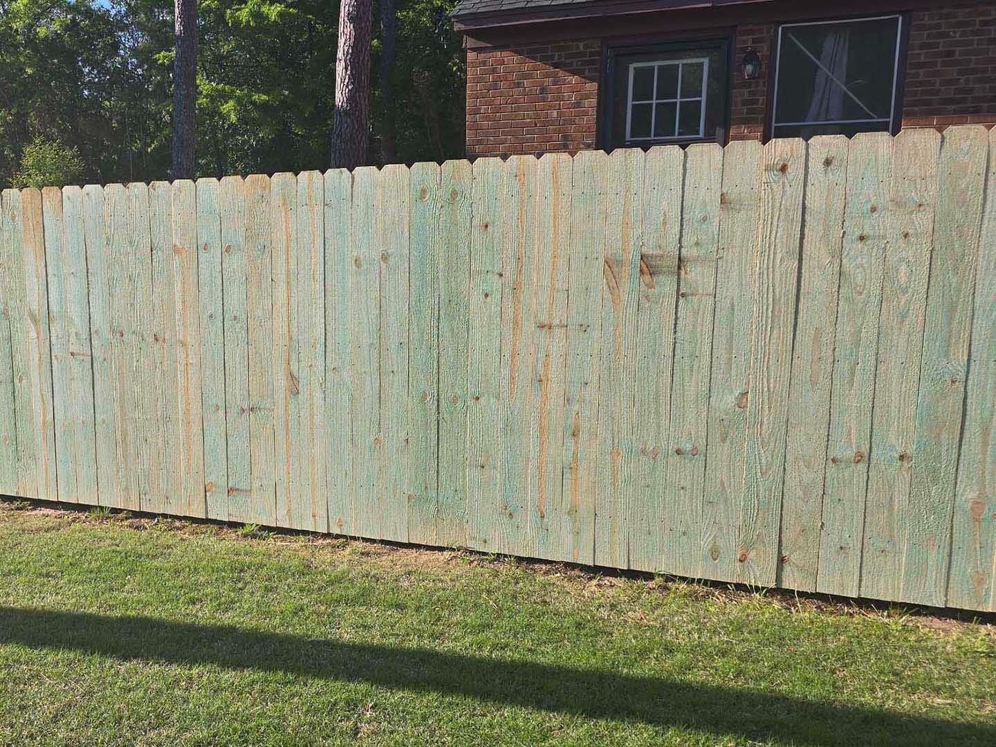A weathered, green-painted wooden fence in front of a brick building and grassy yard.