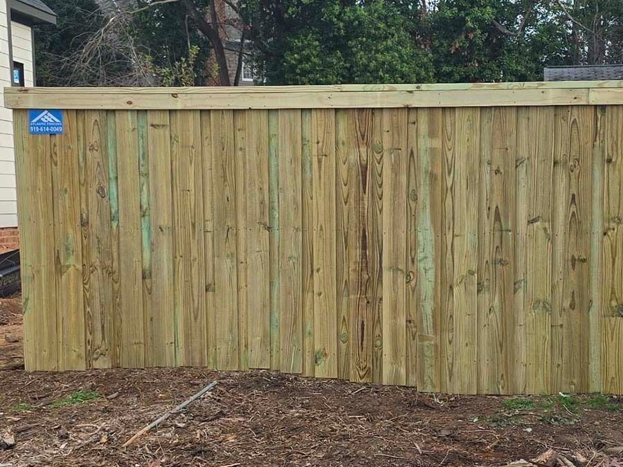 Wooden fence with vertical planks, green-tinged, against a backdrop of green trees.