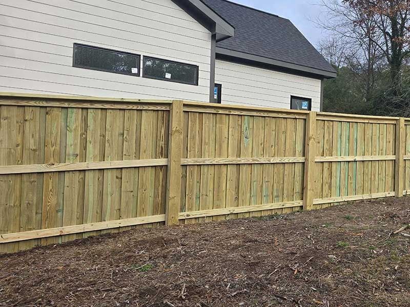 Wooden fence bordering a beige house. Fence is made of vertical boards, brown yard.