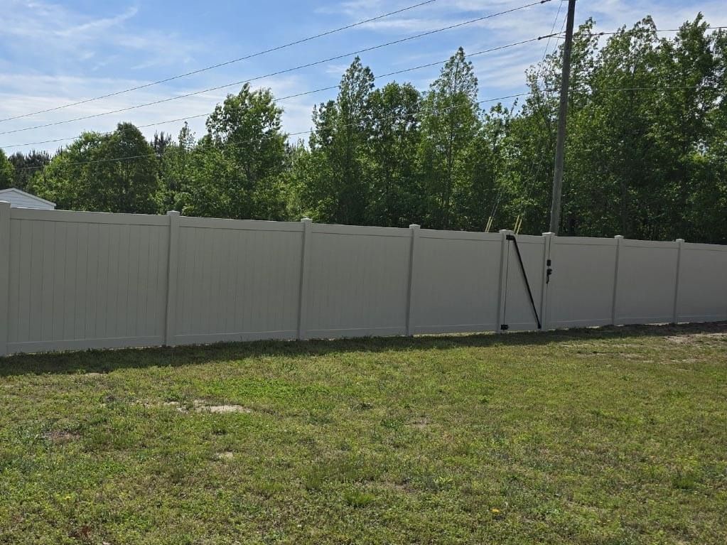 Beige fence in a grassy yard, with trees in the background and a blue sky.