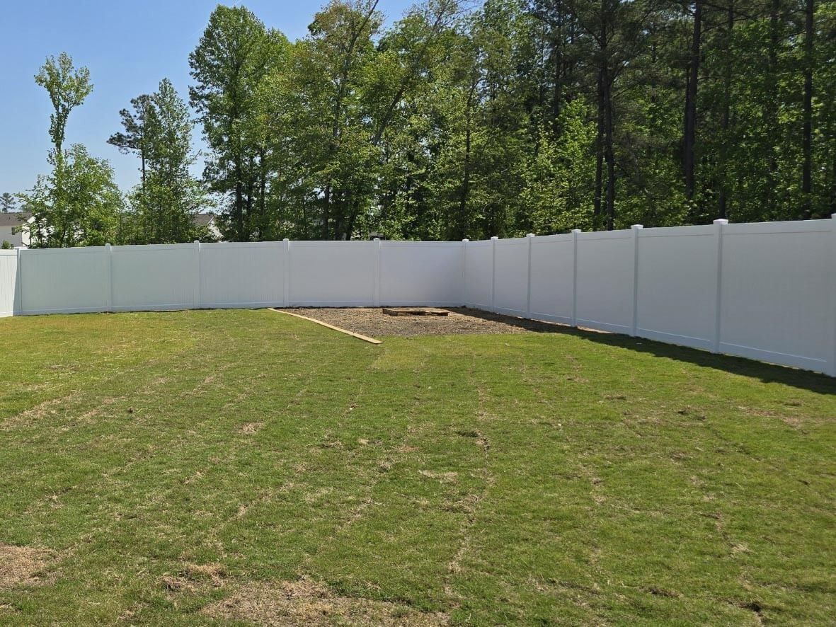 A white fenced backyard with a fire pit surrounded by green grass and trees in the background.