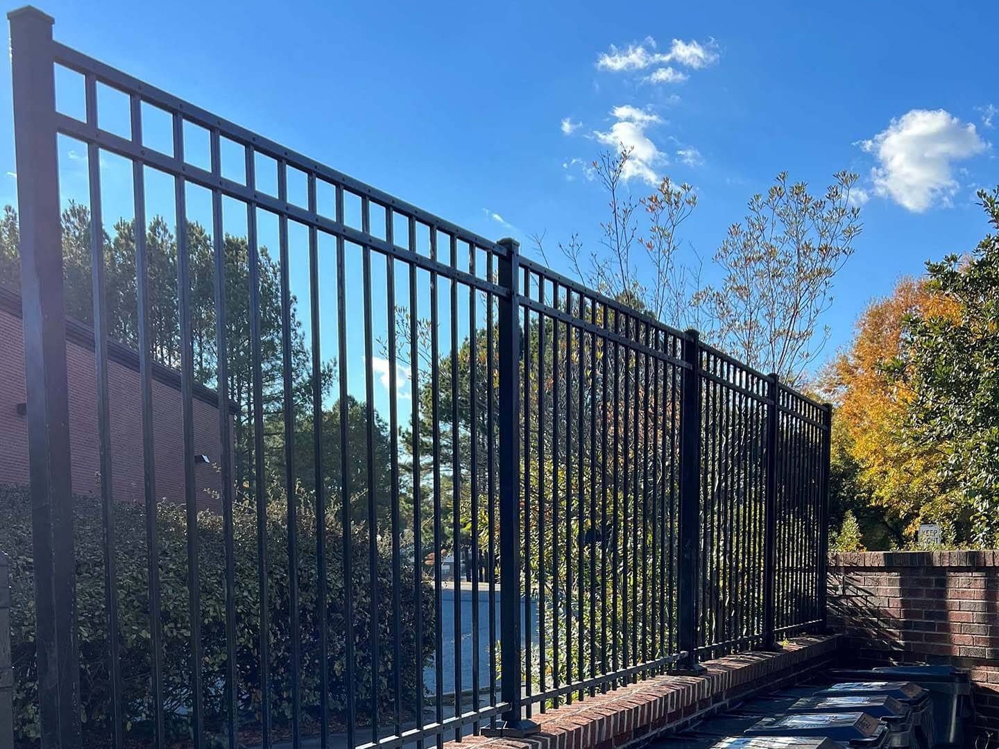 Black metal fence on a brick wall, against a backdrop of trees and blue sky.