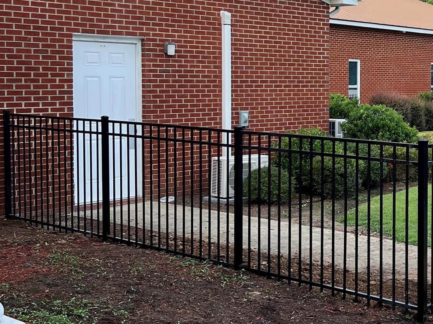 Black metal fence in front of a red brick building with a white door, green bushes, and a concrete pad.