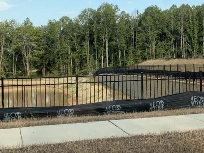 Black fence surrounds a body of water next to a grassy area with trees in the background.