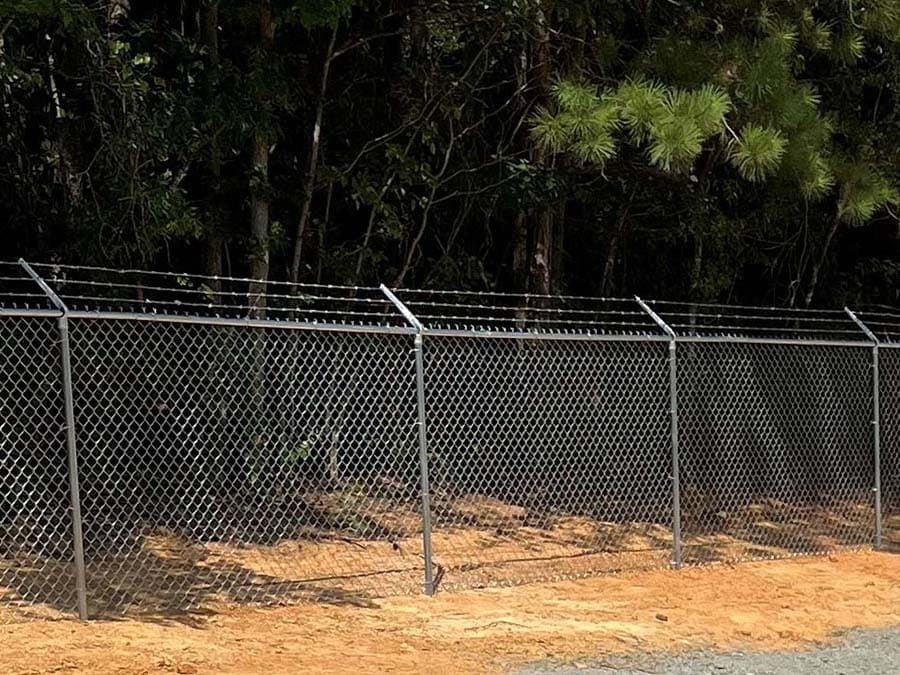 Chain-link fence topped with barbed wire, set against a background of trees and dirt.