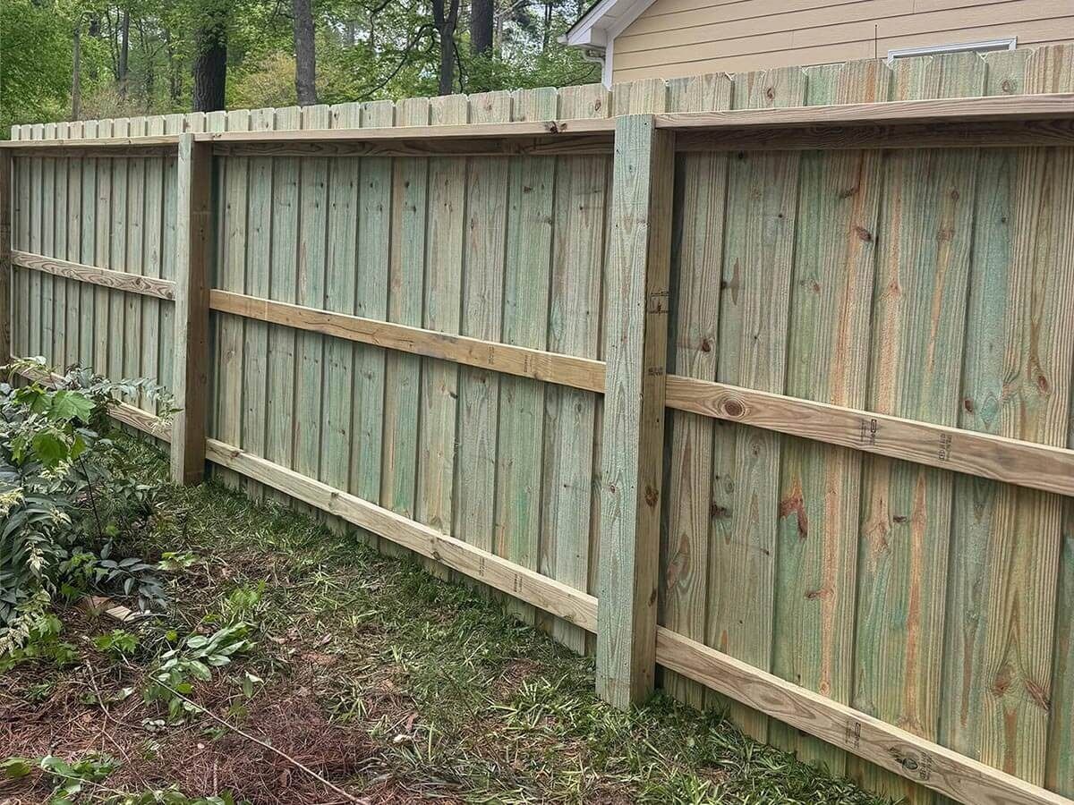 Wooden privacy fence in a yard, green-tinged wood with horizontal supports, on a grassy area.