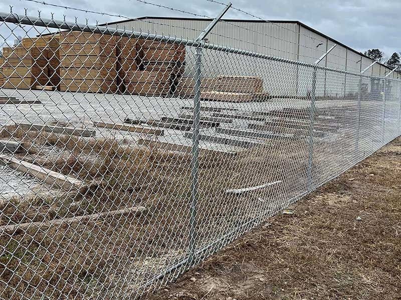 Chain-link fence with barbed wire, enclosing a lumberyard; stacks of wood and a warehouse are in the background.