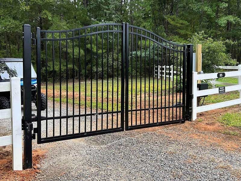 Black metal double gate with curved top, gravel driveway, white fence, and green foliage.